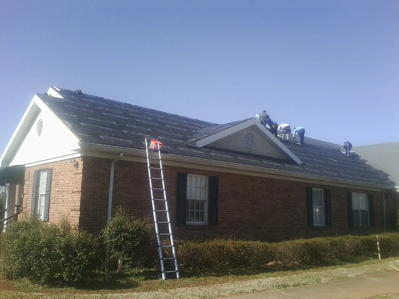 Crew tearing shingles off a brick ranch roof with a ladder set — Emergency Roof Repair work by our crew in Fort Stewart